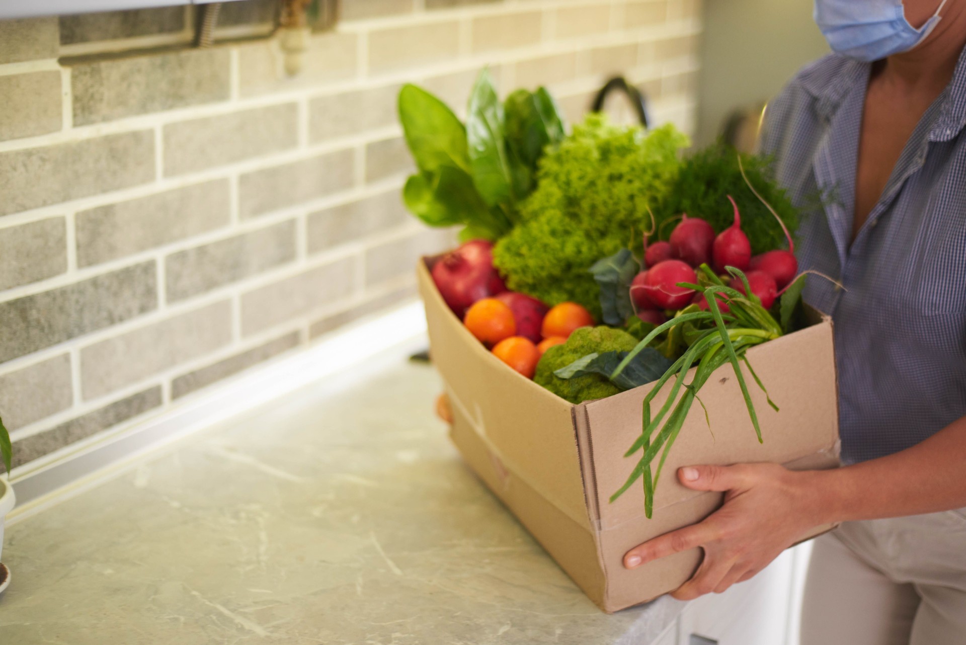 Close-up of woman in medical mask, carrying cardboard box with fresh ripe organic healthy vegetables, fruits and greens